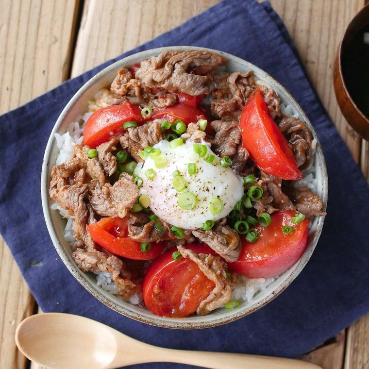 白い皿に盛り付けられた牛こま肉とトマトのスタミナ丼に温泉卵がトッピングされている