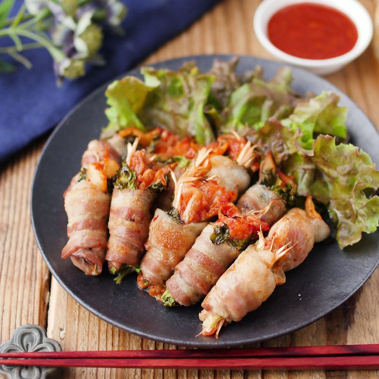 豚バラ肉でキムチを巻いた料理が、黒い皿の上にレタスと一緒に盛り付けられ、そのまわりには赤い箸やたれが置かれている