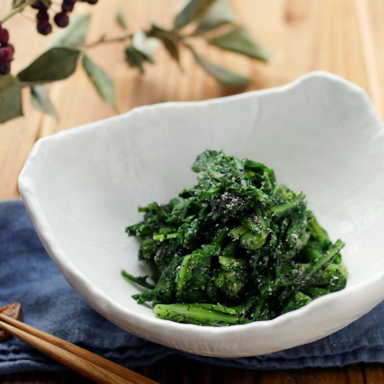 木のテーブルの上に藍色のランチョンマットが敷かれ、その上に白い鉢に盛られた春菊のおひたしと木の箸が置かれている