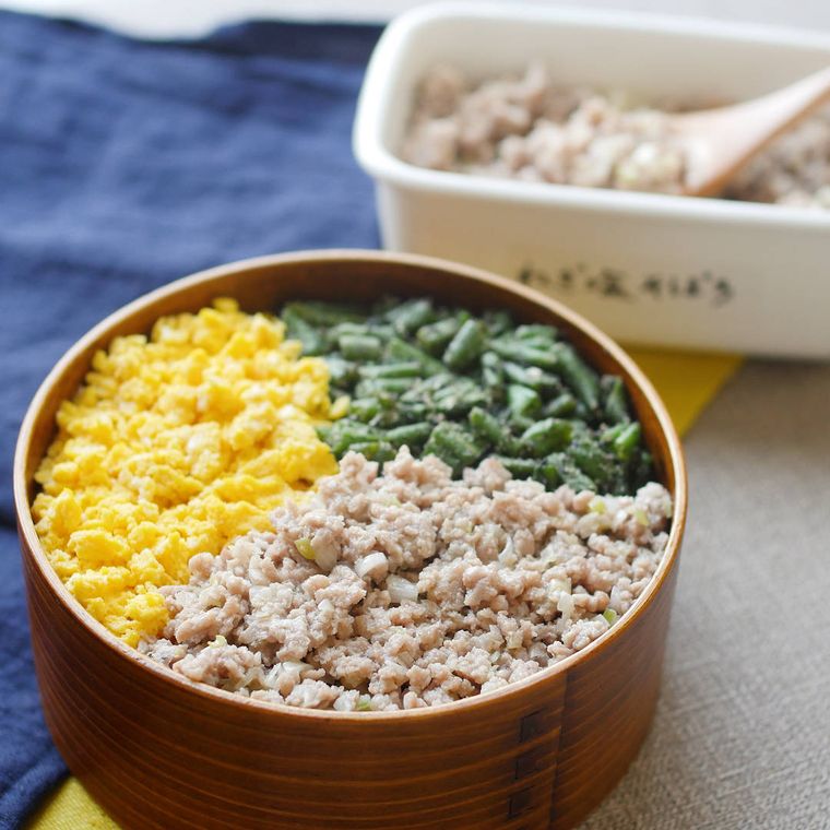 鶏そぼろ、玉子そぼろ、ほうれん草がのった三色丼が曲げわっぱに盛られており、奥には白い容器に入った鶏そぼろが置かれている