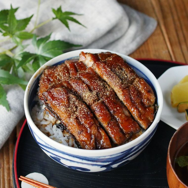 黒いお盆の上に鰻丼とお新香と味噌汁と箸が置かれ、その奥には白い布と緑の植物が置かれている