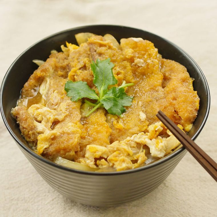 黒い丼にカツ丼が盛られ、その上には三つ葉がのせられており、丼には茶色い箸が添えられている