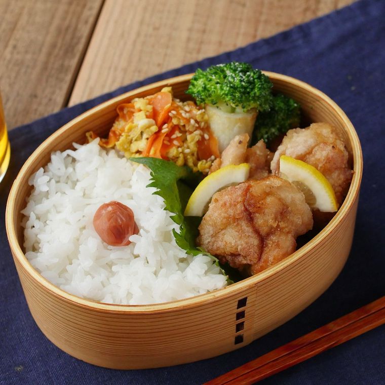 ごはん、梅干し、唐揚げ、大葉などが詰め込まれたわっぱの弁当箱が、青い布の上に置かれている