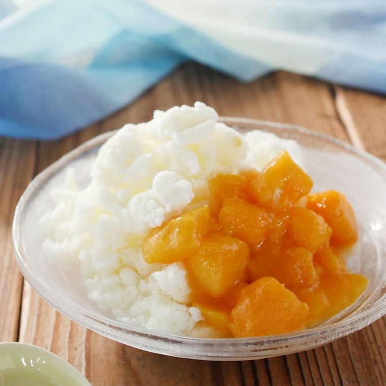 ガラスの器に、果肉入りのマンゴーソースがかかったふわふわのかき氷が盛られており、その奥には青い布が広げられている