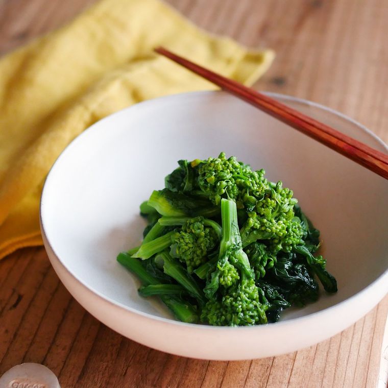 菜の花のおひたしが入った白い器が、木のテーブルの上に置かれており、奥には黄色い布が広げられている