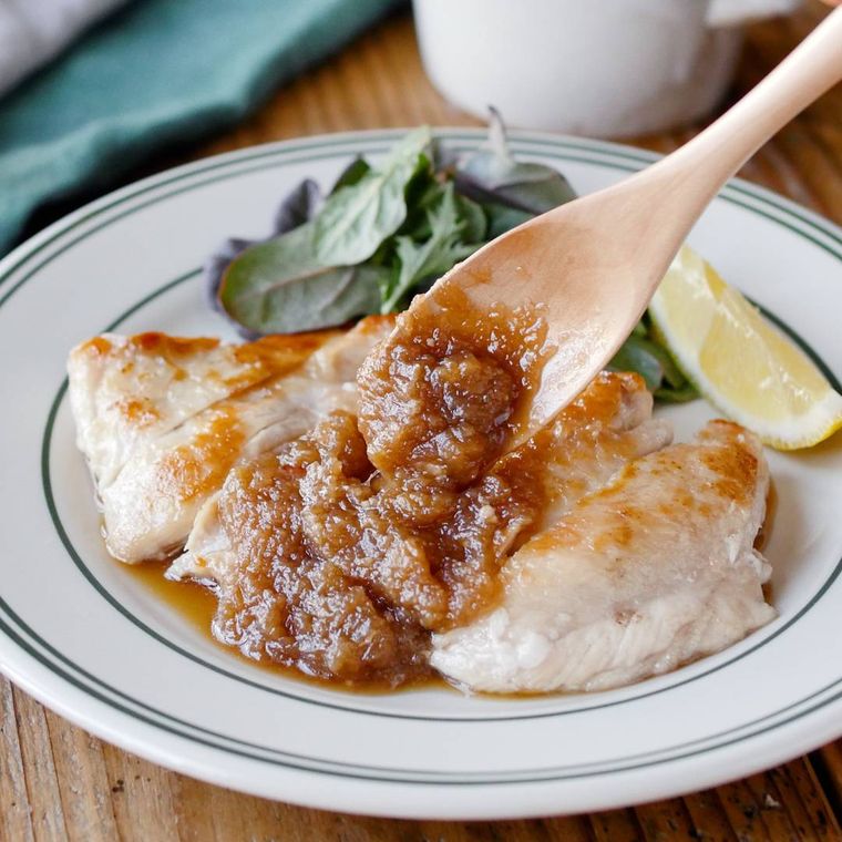 緑の線が入った白い皿に盛り付けられた鶏むね肉のソテーにオニオンソースをかけている様子
