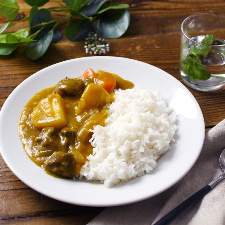 カレーライスが盛られた白い丸皿が、ウッドテーブルの上に置かれており、奥には緑の植物と、グラスが並べられている様子