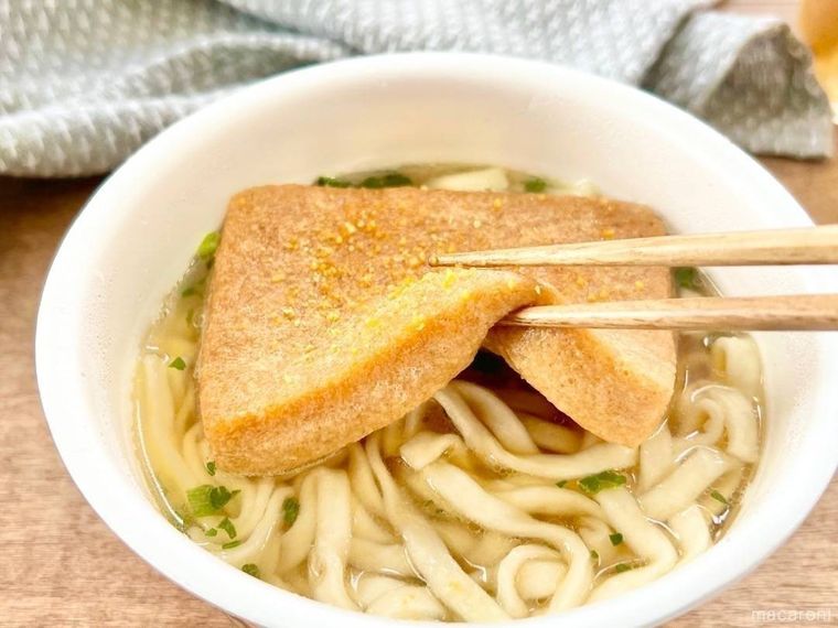 木のテーブルの上に置かれたカップうどんの中から、箸で油揚げを持ち上げようとしており、その奥には灰色の布が広げられている