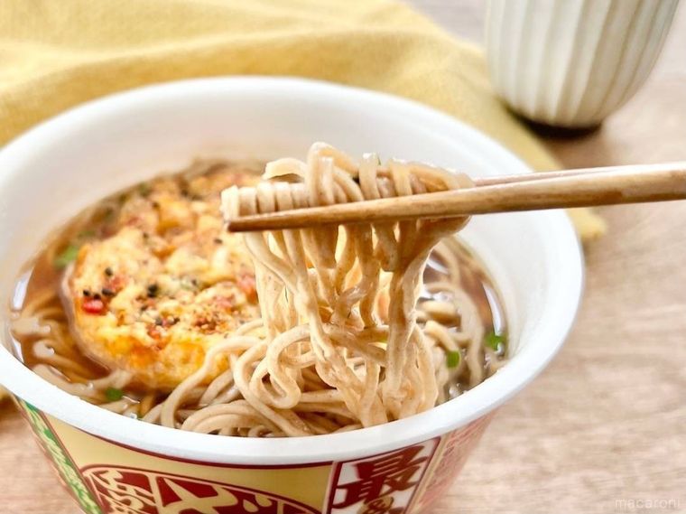 かき揚げがのったカップ麺から、箸でそばを持ち上げており、その奥には黄色の布と、灰色の湯呑みが置かれている