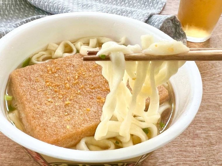 大きな油揚げがのったカップ麺から、箸でうどんを持ち上げており、その奥には灰色の布と、麦茶が入ったグラスが置かれている