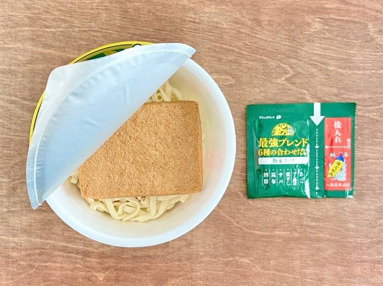 蓋を半分開けたカップ麺の隙間から、乾麺のうどんと油揚げが見えており、その横には、緑色の袋に入った粉末スープが置かれている