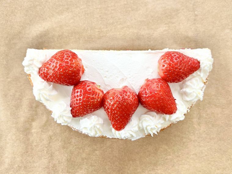 ホイップクリームとイチゴでデコレーションされた半月型のスポンジケーキが、茶色いクッキングシートの上に置かれている