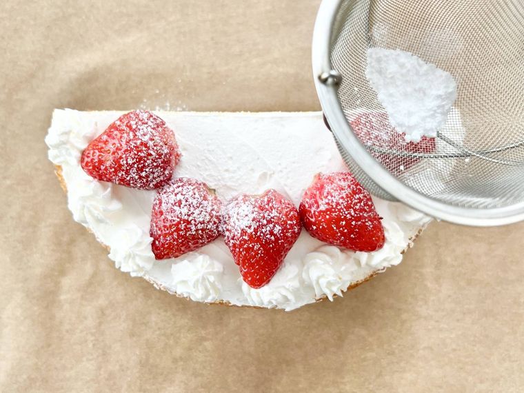 ホイップクリームとイチゴでデコレーションされた半月型のスポンジケーキに、茶こしで粉砂糖をふりかけている