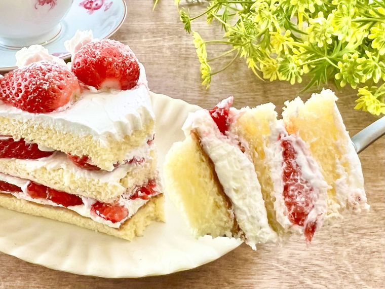 木のテーブルの上に、ショートケーキがのった白い皿が置かれており、フォークでケーキをすくって持ち上げている