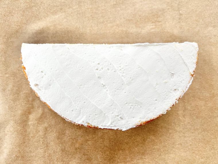 表面にホイップクリームが塗られた半月型のスポンジケーキが、茶色いクッキングシートの上に置かれている