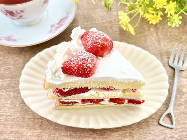 ショートケーキがのった白い皿と、紅茶が入った白いティーカップと、黄色い花とフォークが、木のテーブルの上に並べられている