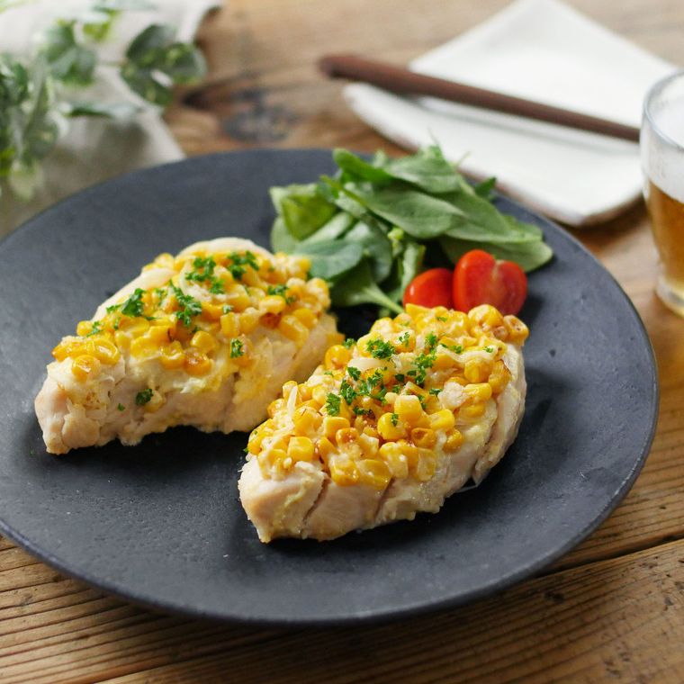 黒い丸皿にサラダチキンのコーンマヨ焼きと付け合わせのサラダが盛られ、机の上に置かれている