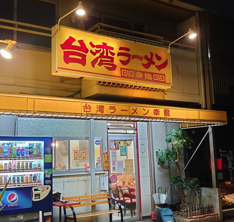 赤文字で屋号が入った黄色い看板 自動販売機とベンチがある外観