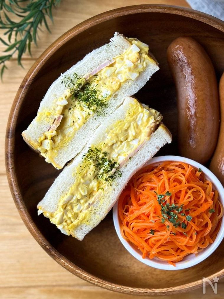 木のトレーにハムたまごサンド、ウインナー、にんじんサラダが盛られている