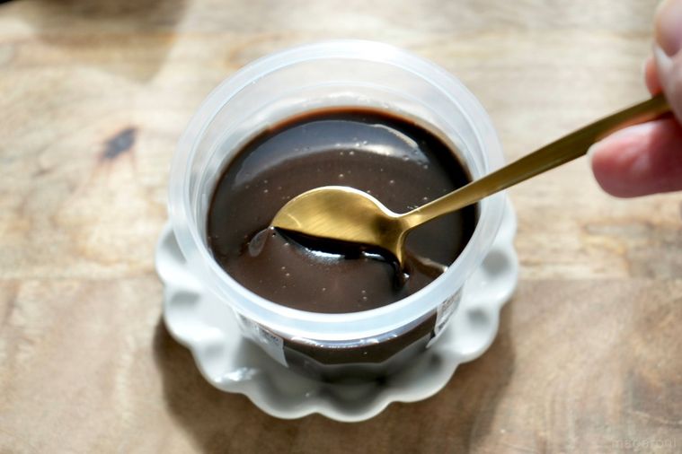 木のカッティングボードと白い小皿にのった黒いショコラプリンを金のスプーンで食べる様子