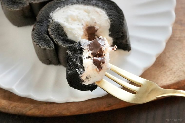 木のカッティングボードと白い皿にのったモノクロのもち食感ロールケーキを金のフォークで食べる様子