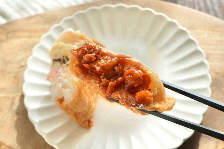 木のカッティングボードにのった白い皿とヤンニョムチキンがのった韓国風いなり寿司を黒い箸で持つ様子