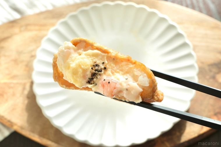 木のカッティングボードにのった白い皿と海老のマヨネーズソース和えがのった韓国風いなり寿司を黒い箸で持つ様子