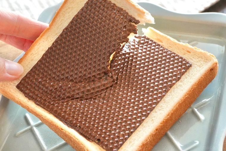 木のカッティングボードとブルーグレーの皿にのったトーストと生チョコスライスを手でちぎる様子