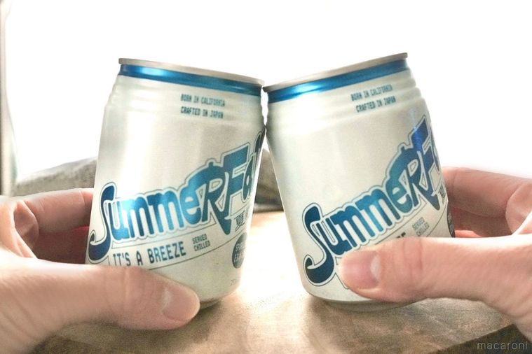 木のカッティングボードにのった白とシルバーと青色のスパークリング日本酒の缶で乾杯する様子