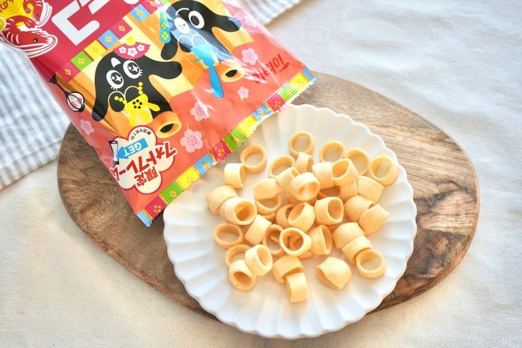 白いテーブルクロスと木のカッティングボードと白い皿にのったポテトスナックとピンク色のパッケージ