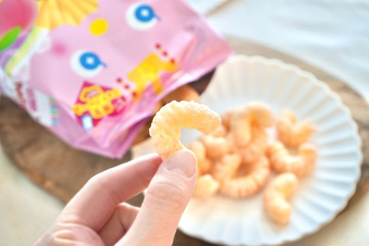 白いテーブルクロスと木のカッティングボードと白い皿にのったひなまつりパッケージの桃味のコーンスナック