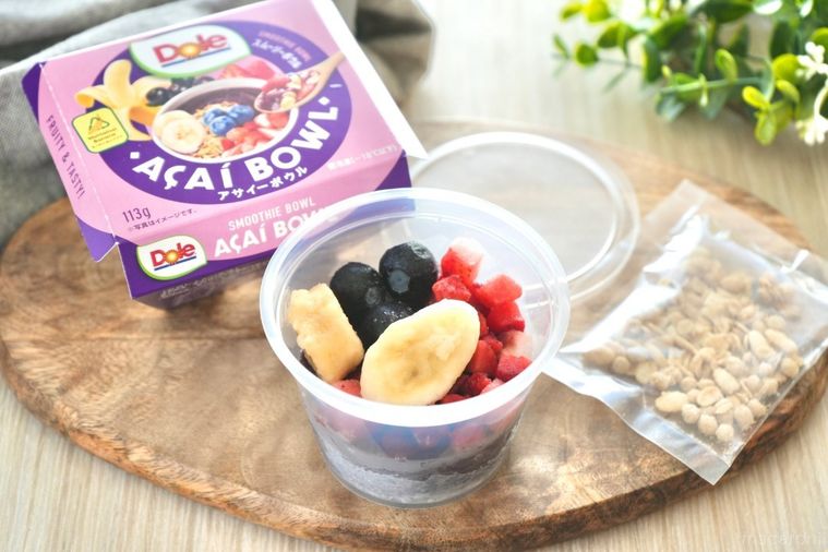 木のカッティングボードにのったカップ入りのアサイーボウルとグラノーラとグリーンとグレーの布