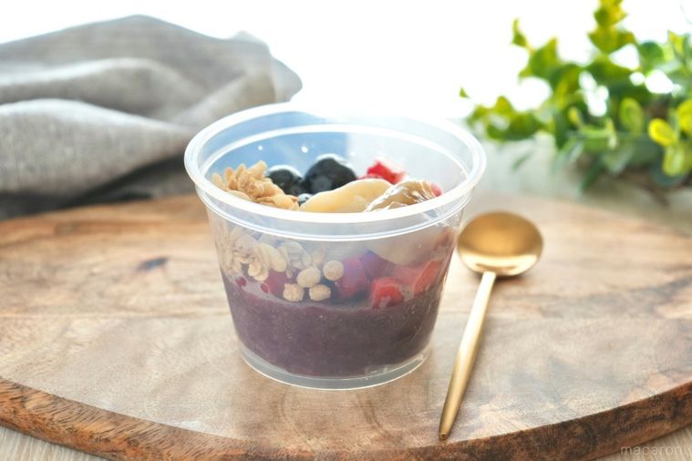 木のカッティングボードにのったプラスチックカップ入りのアサイーボウルと金のスプーンとグリーンとグレーの布