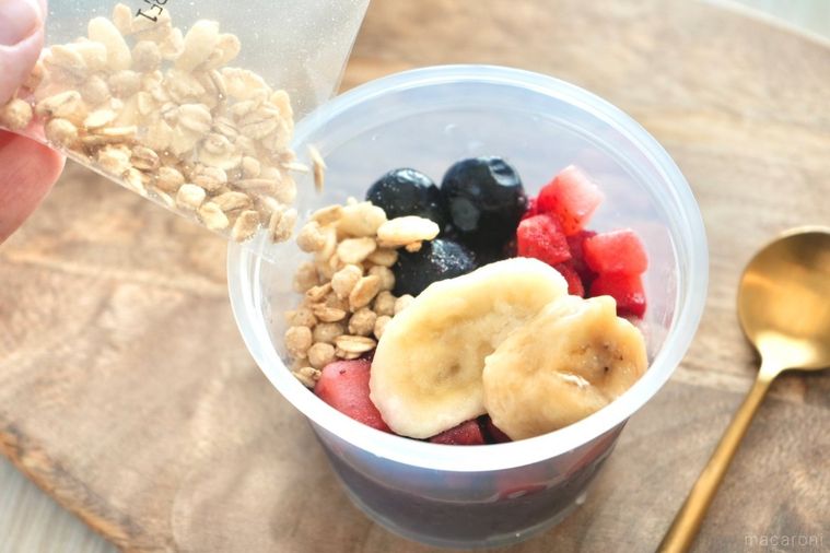 木のカッティングボードにのったプラスチックカップ入りのアサイーボウルにグラノーラをかける様子と金のスプーン
