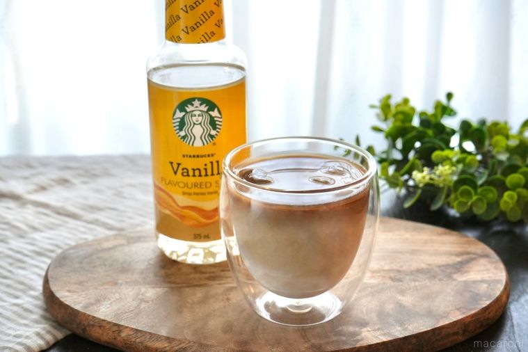 木のカッティングボードにのったグラスにアイスカフェラテバニラフレーバーシロップのボトルとグリーンとストライプの布
