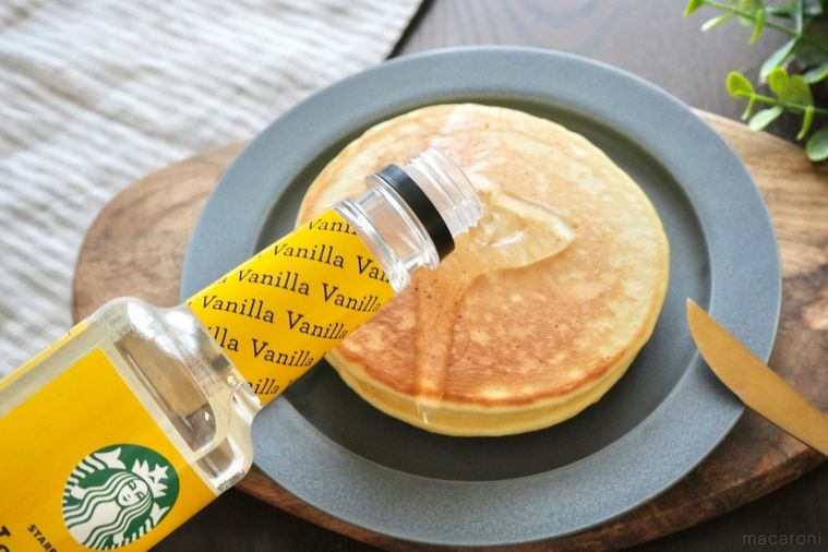 グレーの器にのった2段重ねのパンケーキにシロップをかける様子と金のナイフとフォークとストライプの布