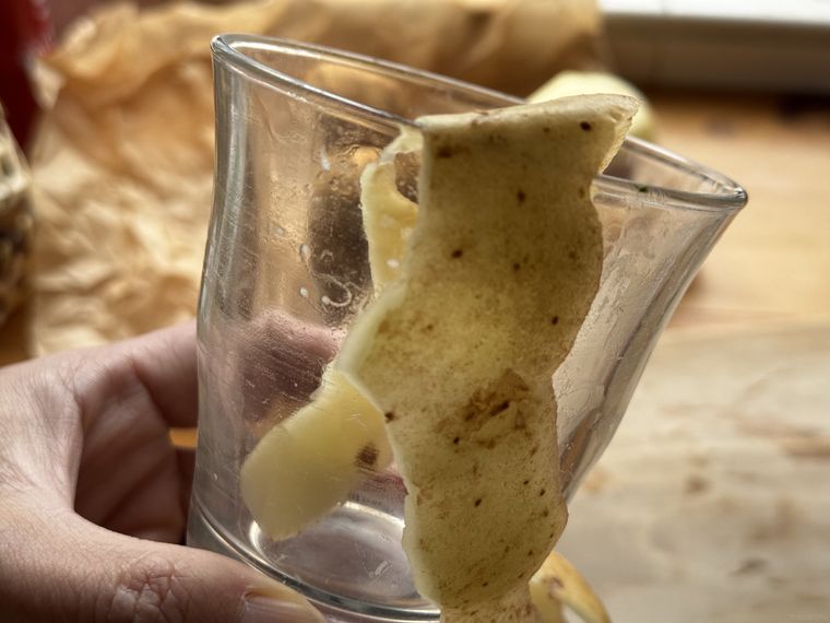 手に持ったガラスコップにじゃがいもの皮が入っており、奥にベージュの包装紙が見えている様子