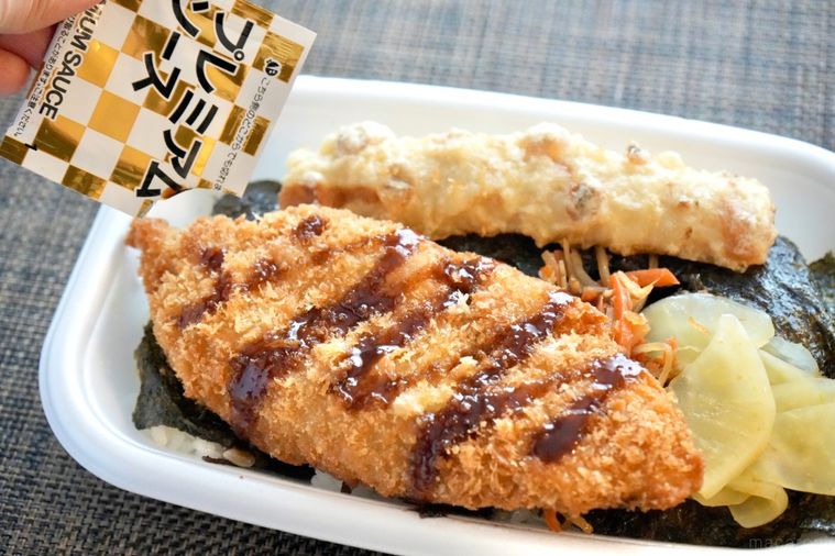 白い容器に入った白身魚のフライとちくわ天ときんぴらごぼうとたくあんがのったのり弁当にソースをかける様子