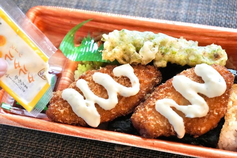 茶色い容器に入った白身魚のフライとちくわの磯辺揚げときんぴらごぼうがのったのり弁当にタルタルソースをかける様子