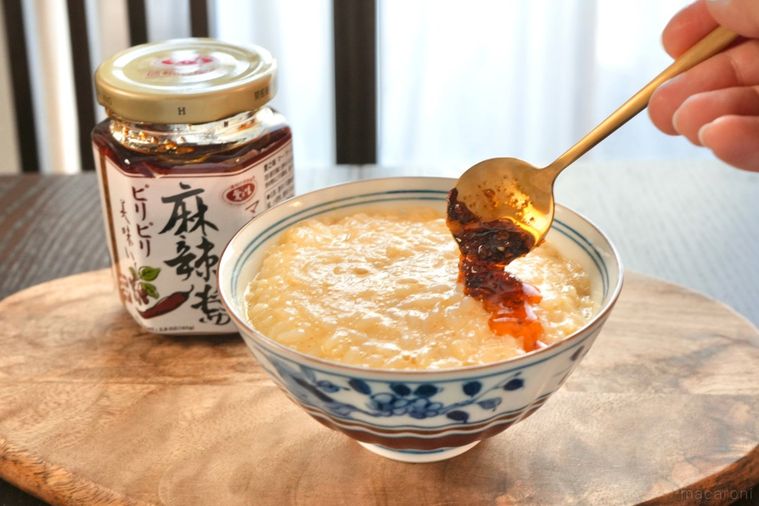 木のカッティングボードにのった茶碗に入った卵かけご飯に金色のスプーンで麻辣醤をかける様子