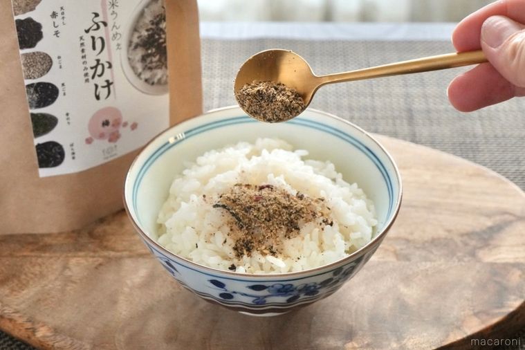 茶色いテーブルクロスと木のカッティングボードにのったごはん茶碗と金のスプーンで米ぬかふりかけを白ごはんにかける様子