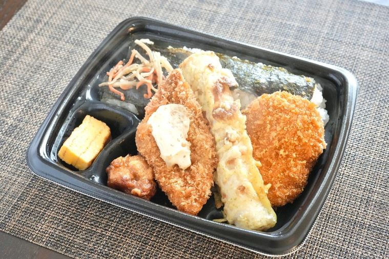 茶色いランチョンマットと黒い容器に盛られた白身魚のフライやちくわの磯辺揚げや揚げ物のせのり弁当