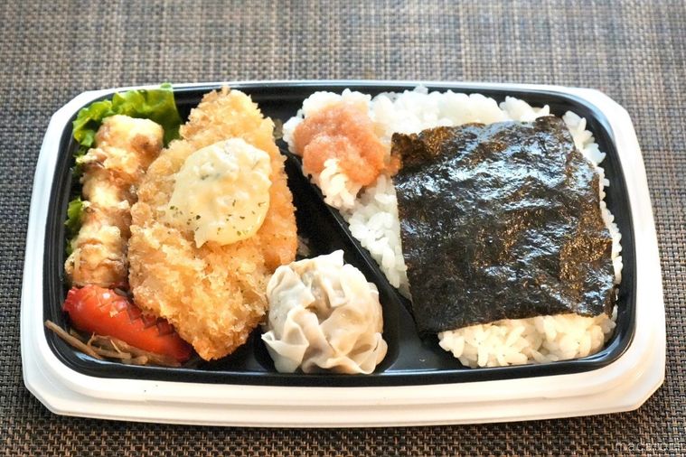 茶色いランチョンマットの上の白身魚のフライとちくわ天と焼売とウインナーと明太子のせのり弁当