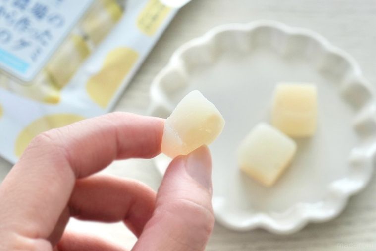 白い木目のテーブルと白い小皿にのった乳白色のキューブ状のグミを手に持つ様子