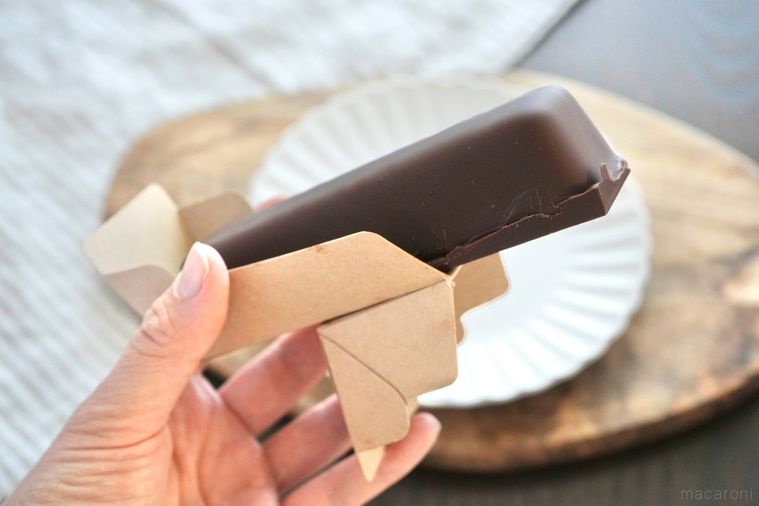 木のカッティングボードと白い皿と紙トレーごとチョコレートスティックケーキを手で持つ様子