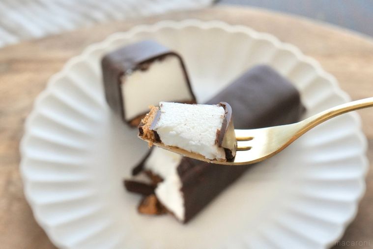 木のカッティングボードと白い皿にのったチョコレートコーティングのムースケーキを金のフォークで食べる様子