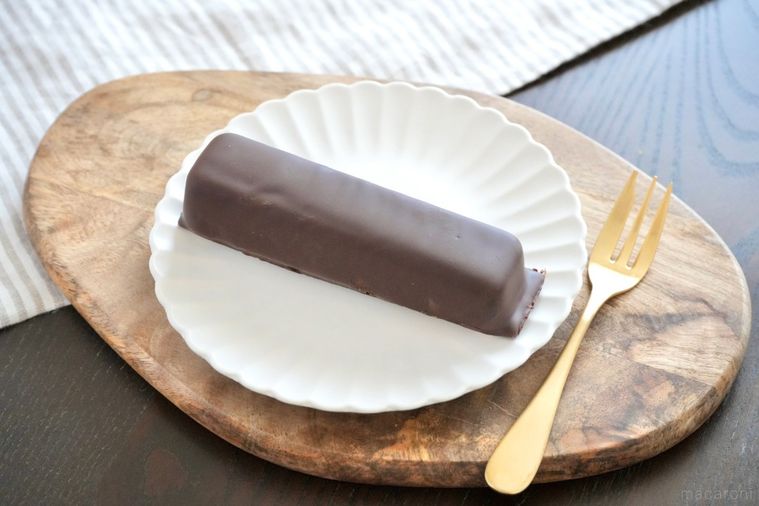 木のカッティングボードと白い皿にのったチョコレートコーティングのムースケーキと金のフォーク