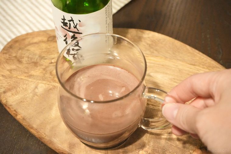 木のカッティングボードにのった日本酒とココア入りのガラスマグを手に持つ様子と緑色の日本酒の瓶