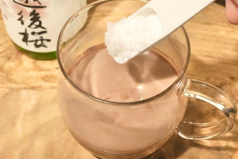 木のカッティングボードにのった日本酒とココア入りのガラスマグに砂糖を入れる様子