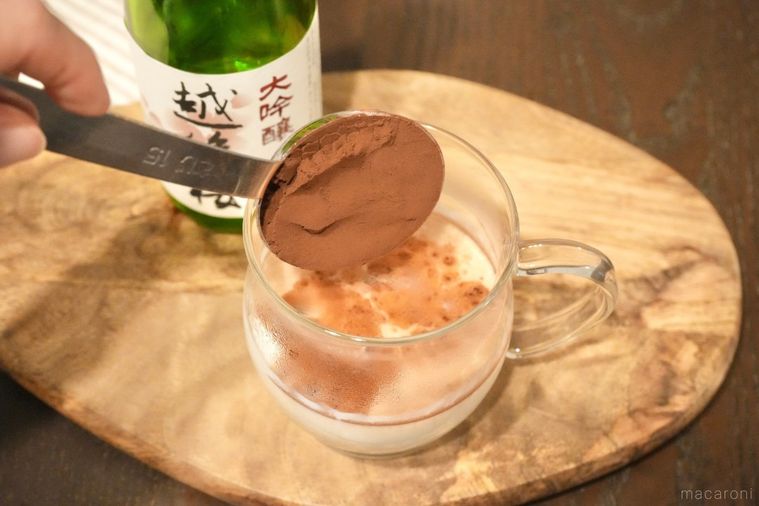 木のカッティングボードにのった日本酒と牛乳入りのガラスマグに純ココアパウダーを計量スプーンで入れる様子と緑色の瓶